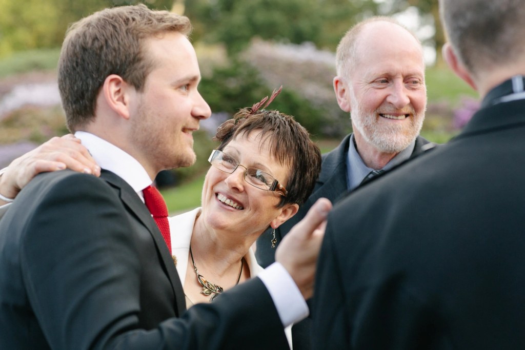 The groom shares a warm moment with his mother and father during the drinks reception in the garden, all three laughing together. Candid documentary wedding photography at the Royal Botanic Garden Edinburgh.