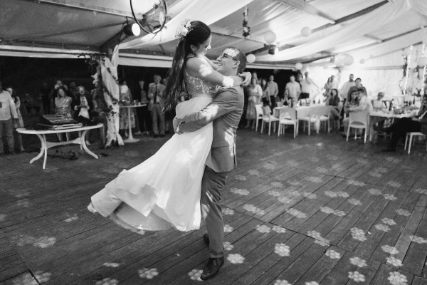 Black and white photo of couple dancing at indoor wedding reception under tent with guests.