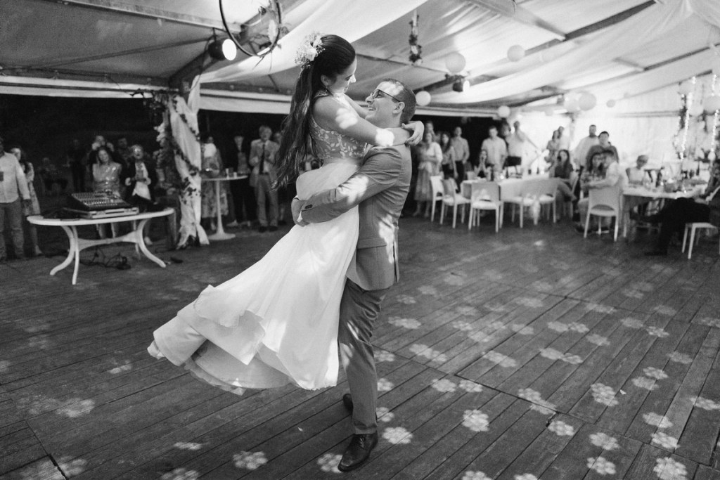 Black and white photo of couple dancing at indoor wedding reception under tent with guests.