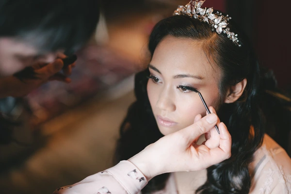 Bride with silver-white floral headpiece focuses on her reflection applying makeup