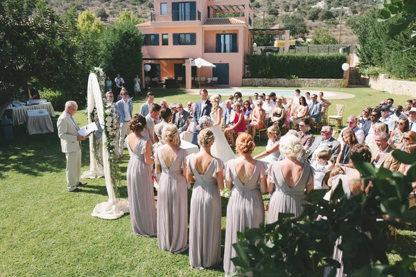 Outdoor ceremony — bride and witnesses facing guests, groom and officiant behind, terracotta house