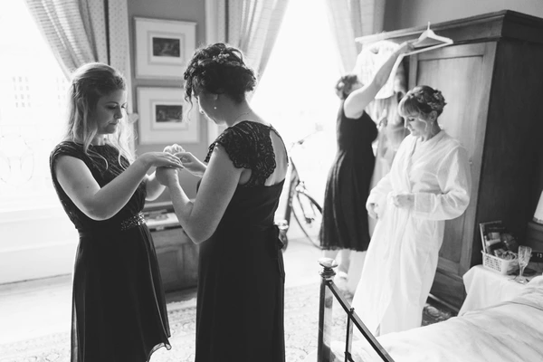 Bride and friends getting ready — woman threads buttons while the white gown waits on the rack