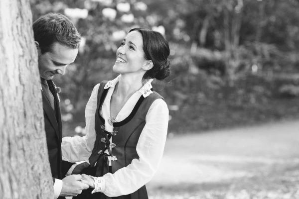 Black and white portrait of the bride in a Norwegian bunad laughing brightly up at the groom beside a tree at the Royal Botanic Garden Edinburgh. Candid documentary wedding photography.