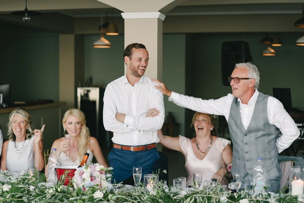 Groom and his father laughing at the dinner, father's hand on the son's shoulder, guests seated