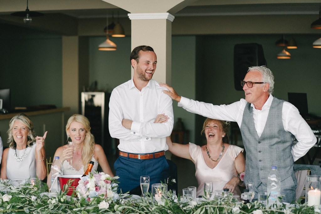 Groom and his father laughing at the dinner, father's hand on the son's shoulder, guests seated