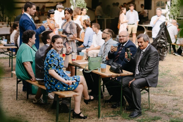 Wedding guests socializing at outdoor reception tables with green and wooden seating during celebration.