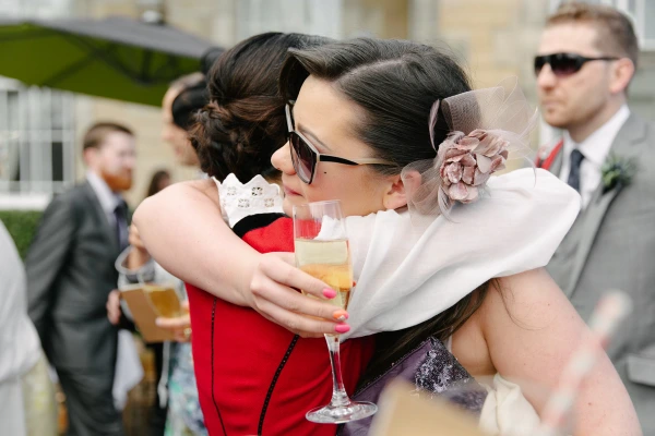 Bride in her Norwegian bunad hugging a guest in a purple sequined dress and sunglasses at the outdoor drinks reception, champagne glass in hand. Candid post-ceremony embrace.