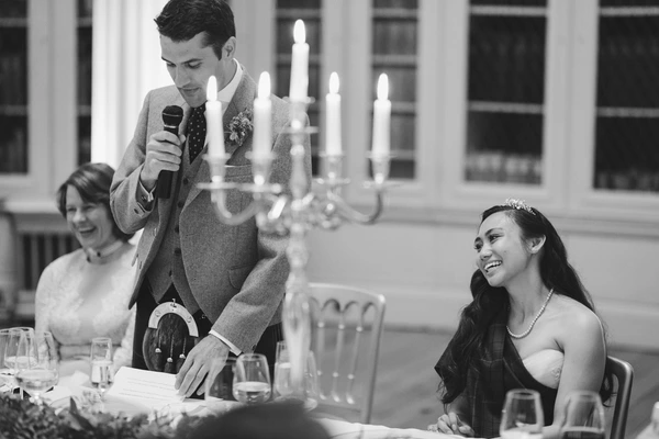 Groom speaks at the wedding dinner with a microphone, bride smiling beside him — candlelit