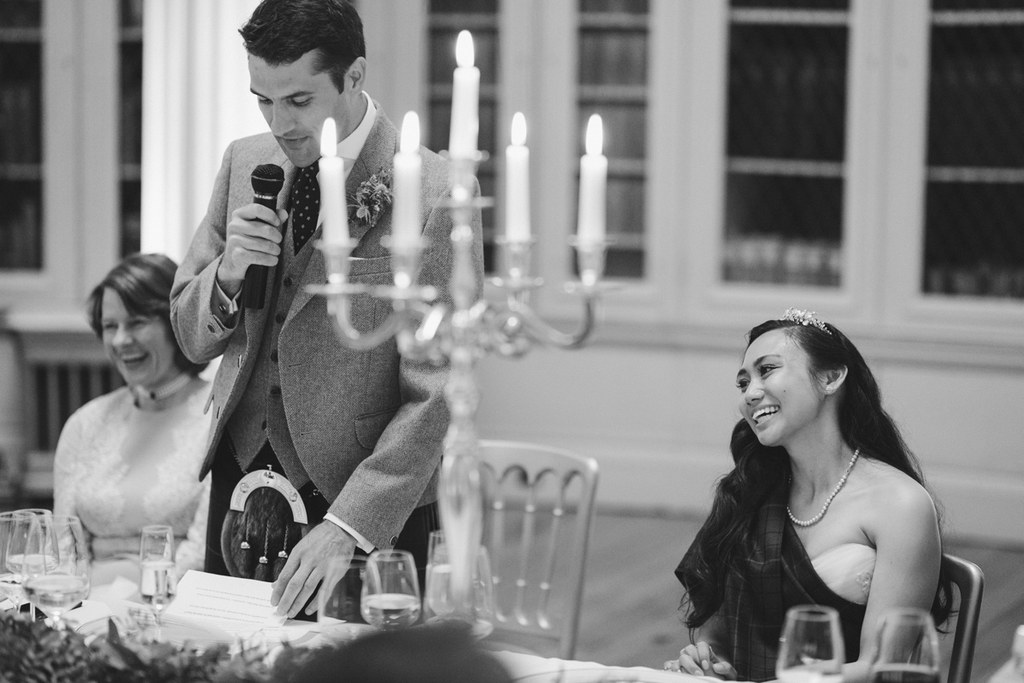 Groom speaks at the wedding dinner with a microphone, bride smiling beside him — candlelit