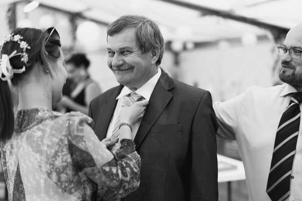 Black and white candid photo of a woman adjusting an older man's tie at a wedding. Emotional guest moment.