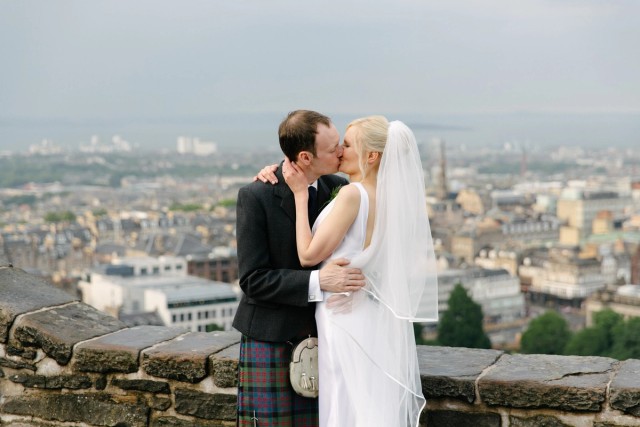 Bride and groom exchanging a kiss while standing on the wall of the Edinburgh Castle with New Town and the harbour behind them as the backdrop