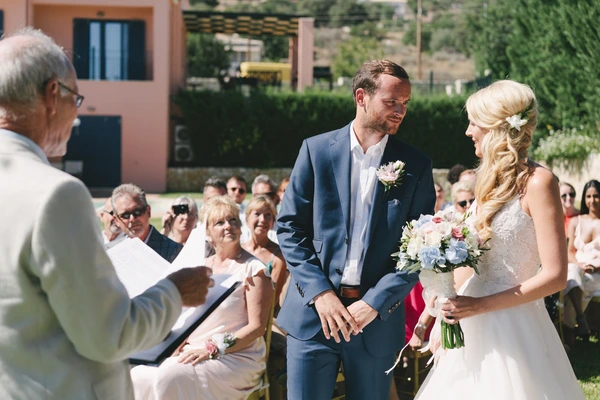 Groom in blue suit and bride in white look at each other at the ceremony — sunny garden, guests seated
