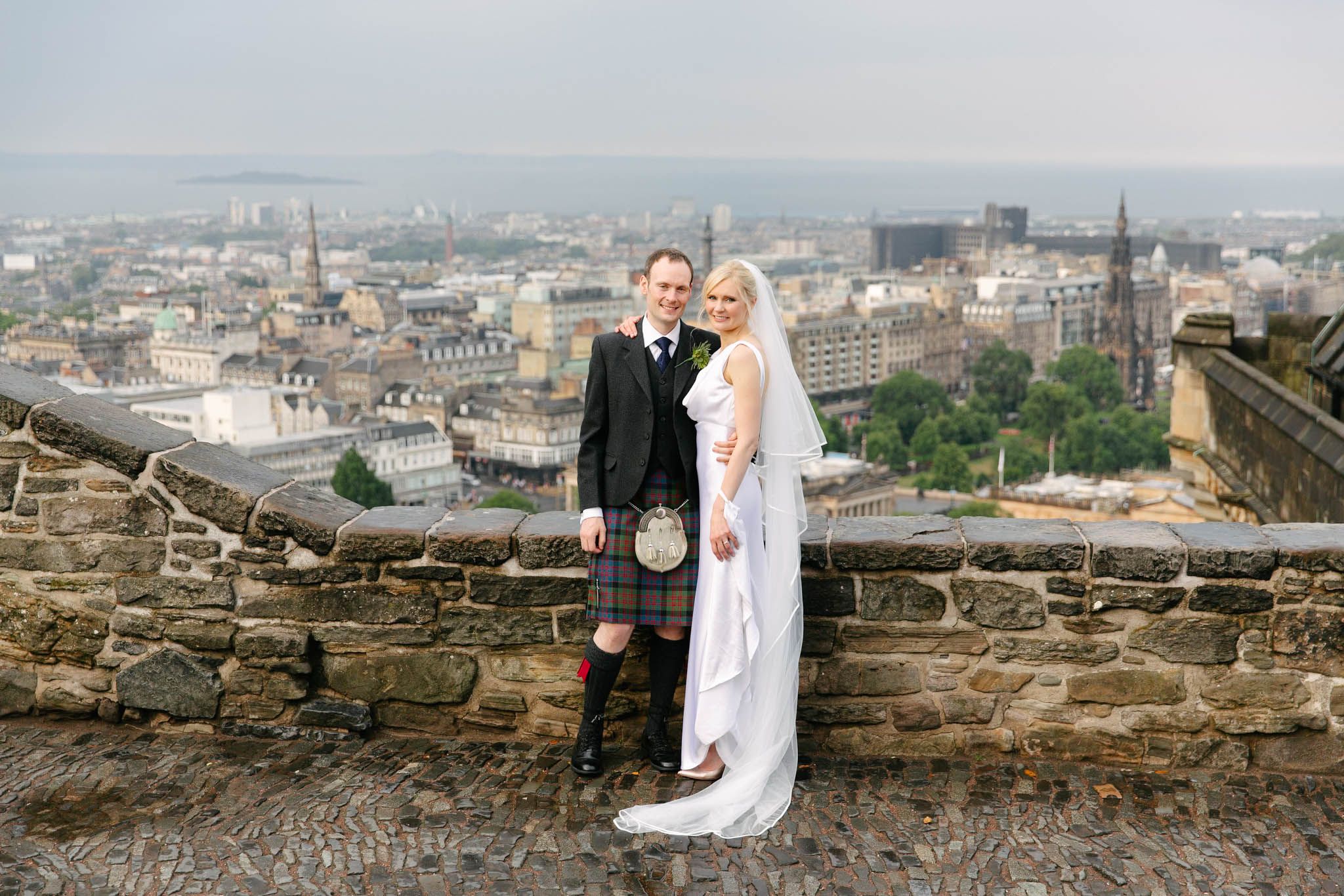 Bride and groom in Scottish attire posing at stone wall overlooking Edinburgh cityscape on their wedding day.