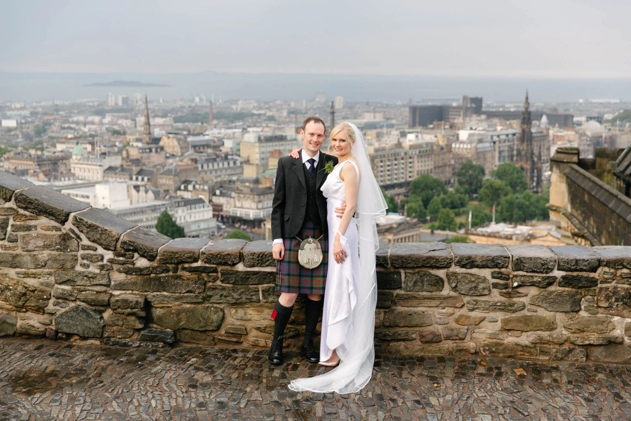 Bride and groom in Scottish attire posing at stone wall overlooking Edinburgh cityscape on their wedding day.
