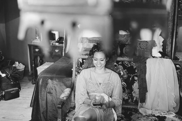 Bride smiles in a chair looking at her phone during preparations — black-and-white
