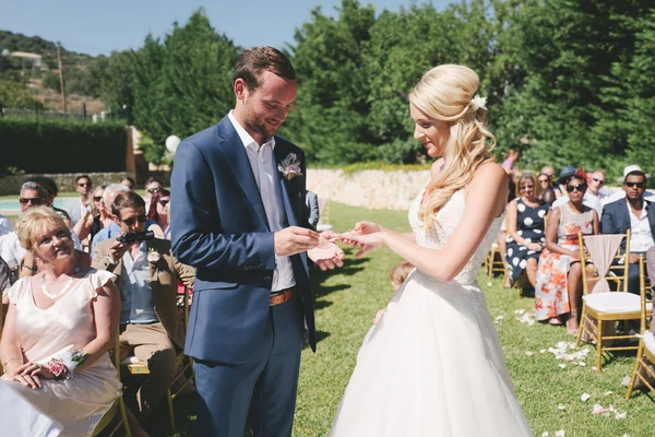 Groom in blue suit places ring on bride's white lace hand at the outdoor ceremony — guests smiling
