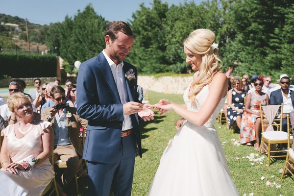 Groom in blue suit places ring on bride's white lace hand at the outdoor ceremony — guests smiling