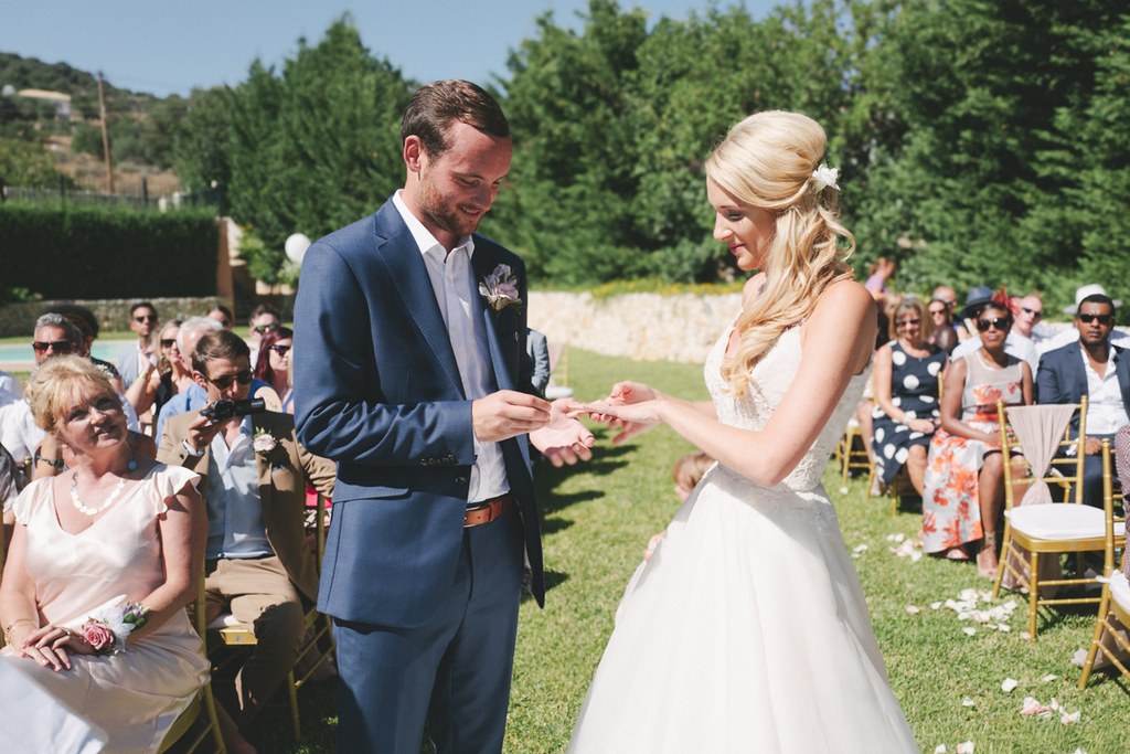 Groom in blue suit places ring on bride's white lace hand at the outdoor ceremony — guests smiling
