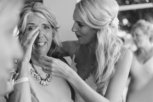 Touching moment — bride gently wipes tears from her mother's face, jewels and rings visible. B&W.