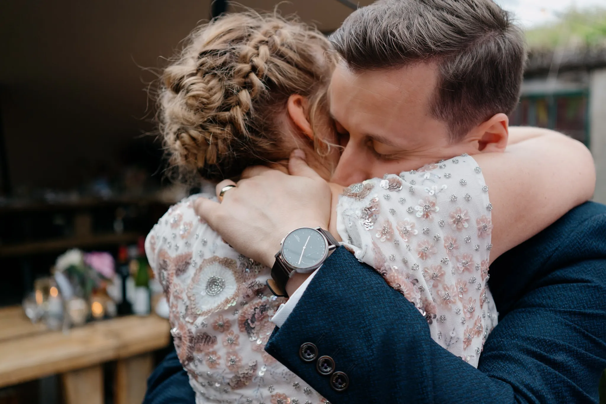 Bride and groom share emotional embrace, her braided updo and beaded dress visible, his navy suit and watch prominent.