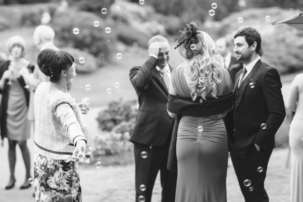 Black and white image of soap bubbles floating among wedding guests outdoors at the Royal Botanic Garden Edinburgh, with a blonde woman in a fascinator at the centre. Candid documentary wedding photograph.