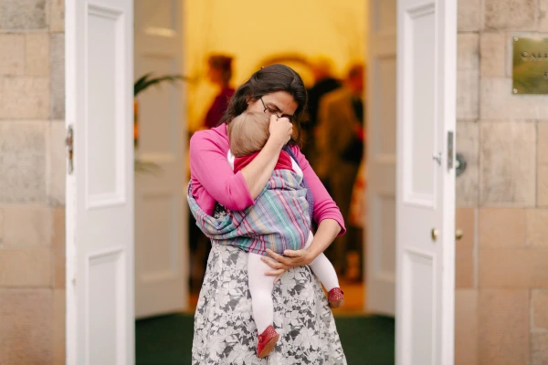A woman in a pink top carries and nuzzles a baby in a colourful wrap carrier in the doorway of the wedding venue, framed by white double doors. Candid documentary wedding photograph.