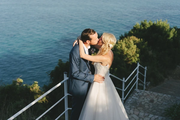 Bride and groom kiss at a stone wall overlooking the sea, surrounded by green vegetation