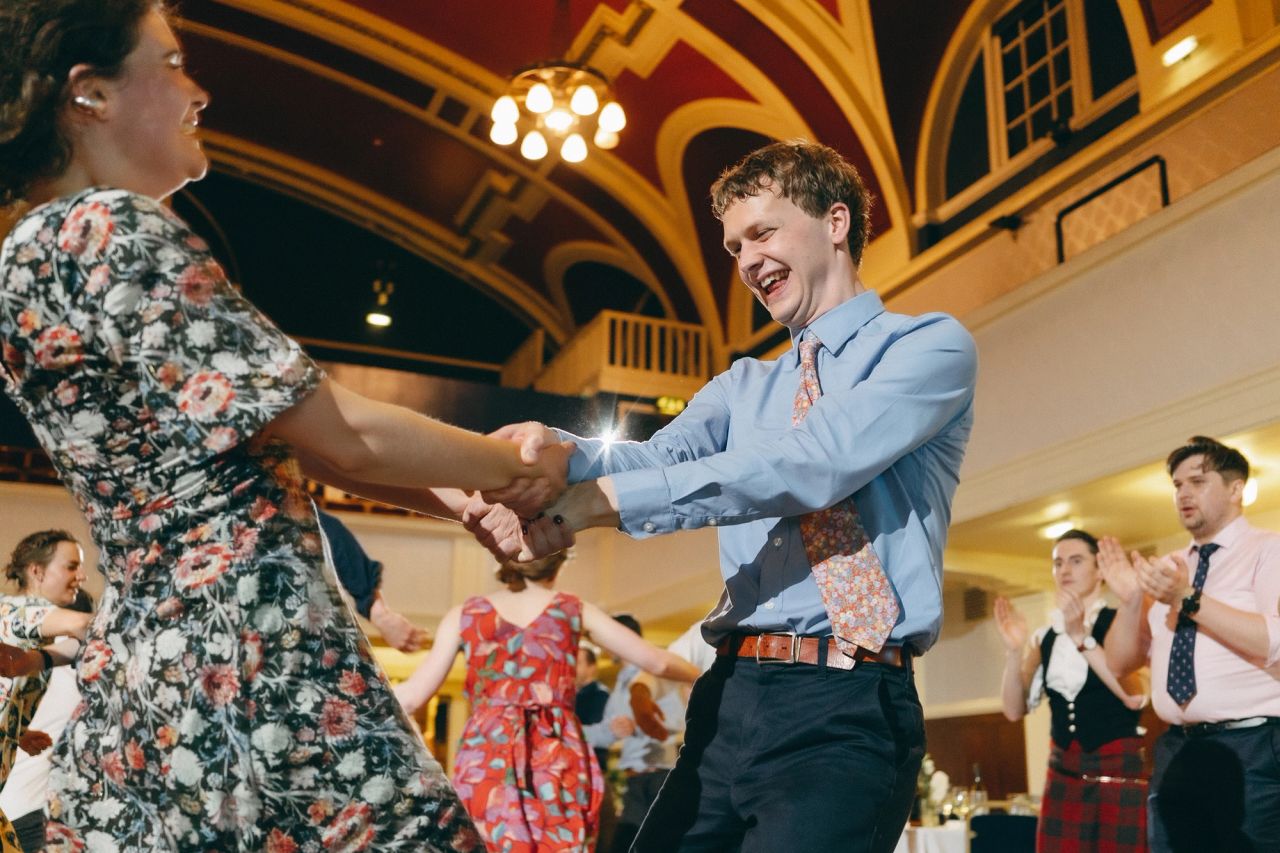 Vidám esküvői tánc egy elegáns, aranyozott ballroomban. Fiatalok színes ruhákban táncnak és mosoly közben kézenfogva ünnepelnek.
