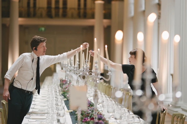 Wedding coordinators light candles among red flowers at the long table in an elegant hall