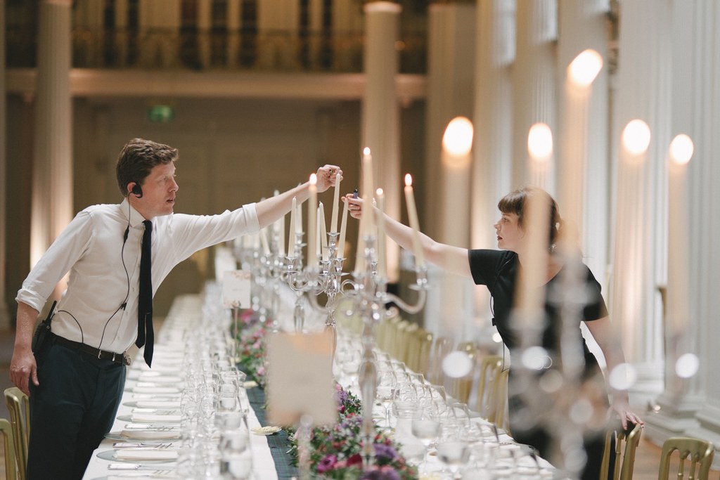 Wedding coordinators light candles among red flowers at the long table in an elegant hall