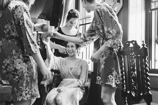 Bride laughing in a chair, guests in floral kimonos playfully draped around her — sunlit elegant room