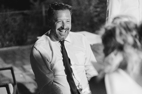 Groom in white shirt and dark tie laughing and talking with guests — warm evening mood