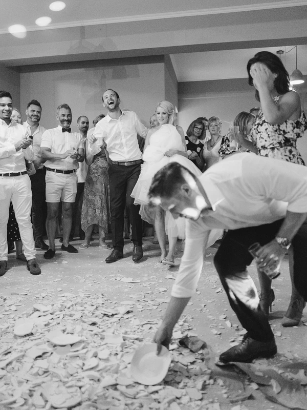Guests circle the groom who shouts with joy, bride covers her mouth in surprise — confetti flying indoors