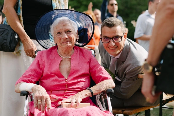 Elderly woman in pink dress and navy hat smiling with groom at outdoor wedding celebration event.