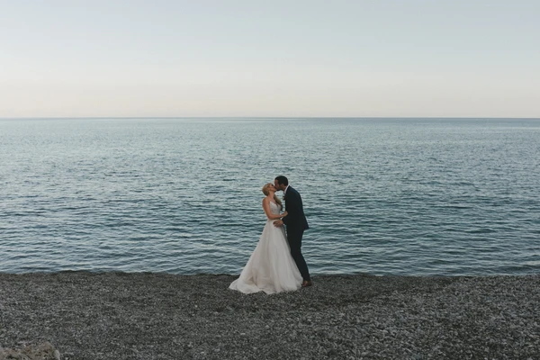Bride and groom kiss on the beach — white pearlescent dress, black suit, turquoise sea and clear sky