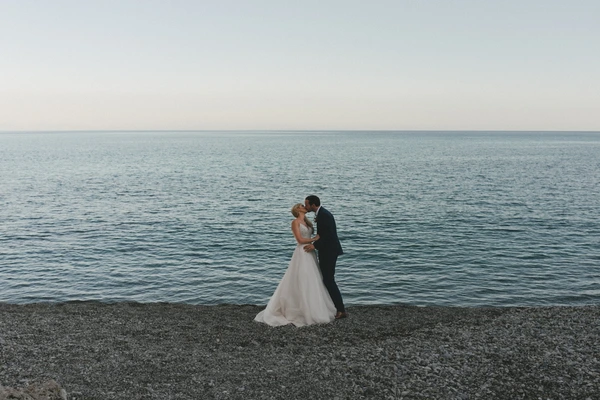 Bride and groom kiss on the beach — white pearlescent dress, black suit, turquoise sea and clear sky