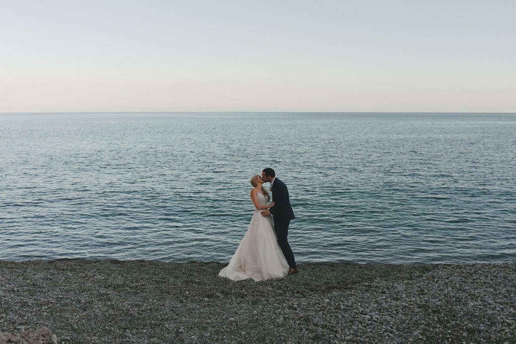Bride and groom kiss on the beach — white pearlescent dress, black suit, turquoise sea and clear sky