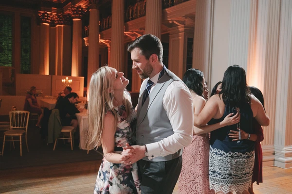 Groom in white shirt and waistcoat dances with bride in floral dress under orange light