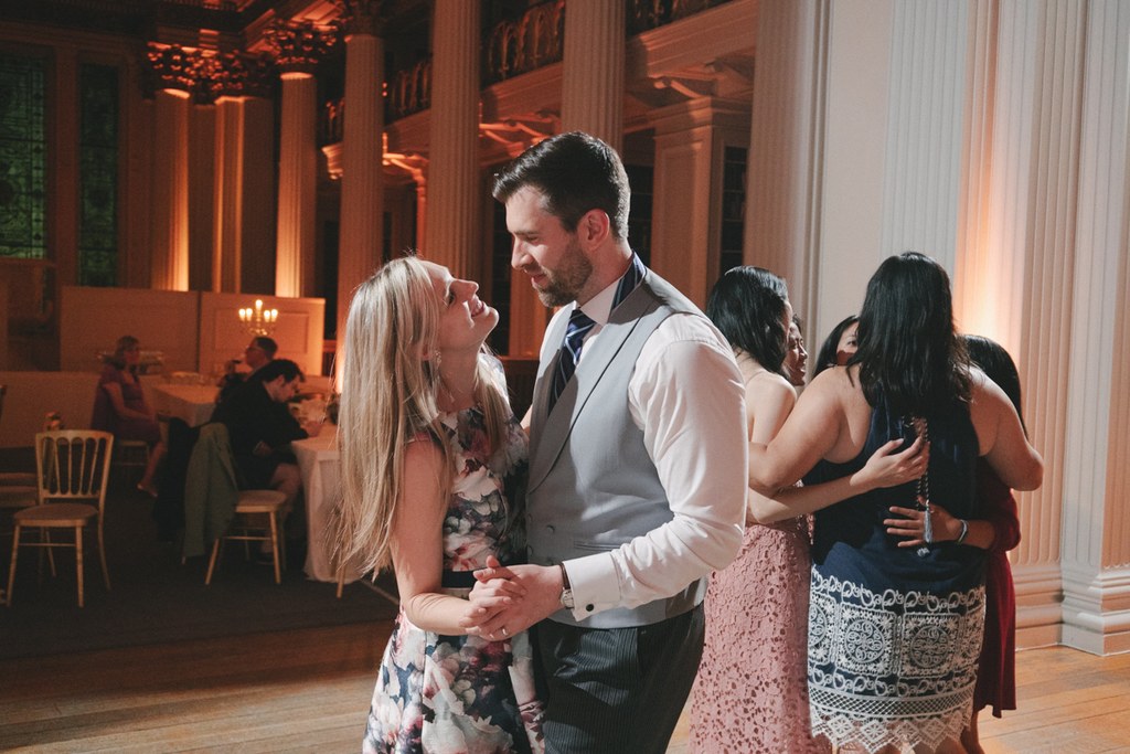 Groom in white shirt and waistcoat dances with bride in floral dress under orange light