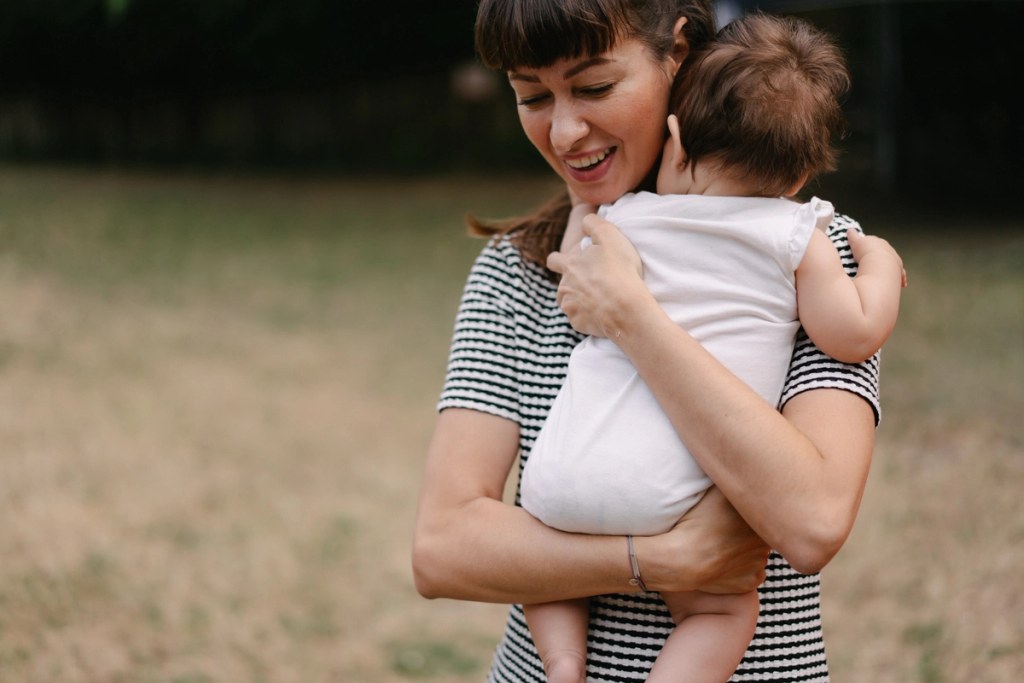 Woman holding and hugging toddler joyfully at outdoor wedding in striped shirt with green background.
