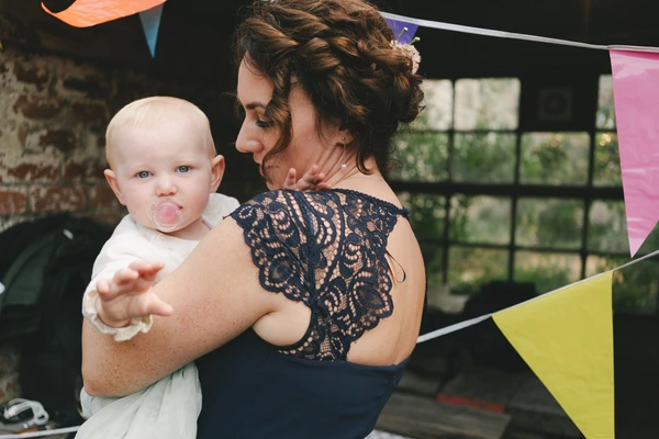 Woman in black lace holds a blonde little boy — bunting, colourful flags, old brick background