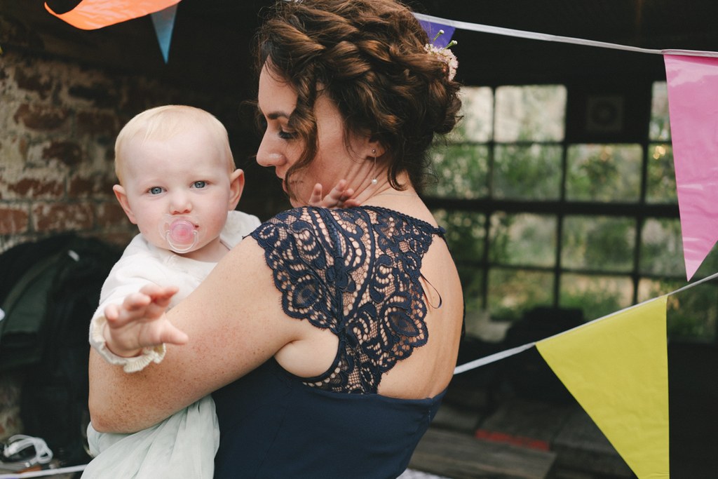 Woman in black lace holds a blonde little boy — bunting, colourful flags, old brick background