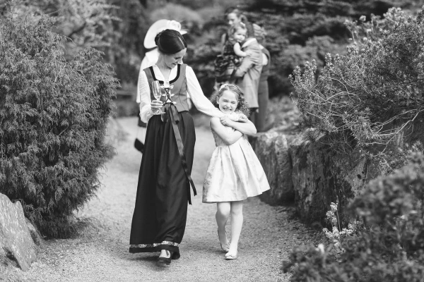 Black and white image of a woman in a Norwegian bunad walking hand in hand with a smiling flower girl along a garden path, with another woman carrying a toddler in the background. Documentary wedding photography.