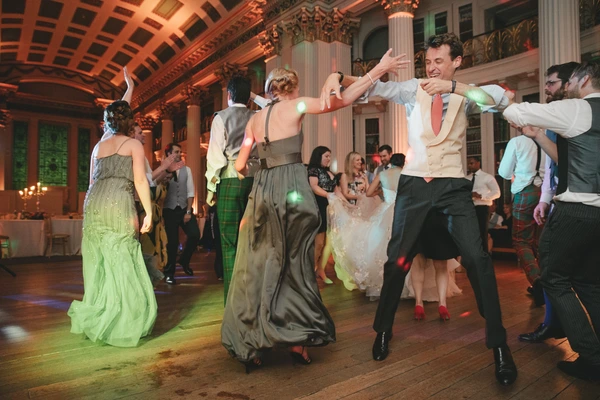 Groom and guests dance in the ornate hall — warm orange light, man in white waistcoat and pink tie