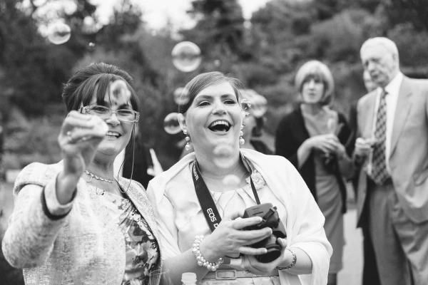 Black and white image of two women laughing and reaching for soap bubbles at the wedding drinks reception, with other guests watching in the background. Candid documentary wedding moment.