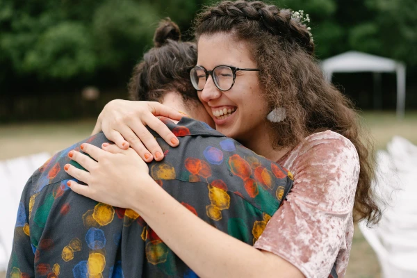 Woman with glasses hugging guest in patterned shirt at outdoor wedding celebration with joyful expression.