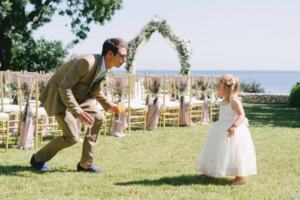 Groom playfully leans toward the little flower girl at the ceremony venue, flowers and chairs around
