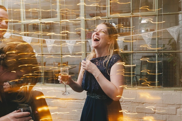 Girl in dark blue dress holds a glass of champagne, laughing amid golden fairy lights