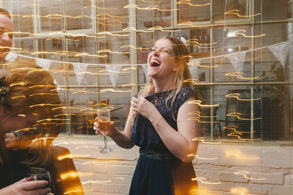 Girl in dark blue dress holds a glass of champagne, laughing amid golden fairy lights