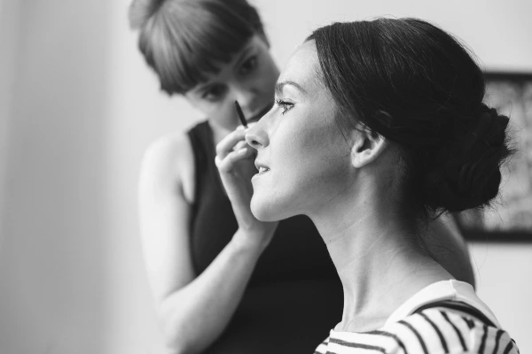 Black and white close-up of a make-up artist applying eyeliner to the bride in profile, bride's hair in an elegant updo. Candid bridal preparations.